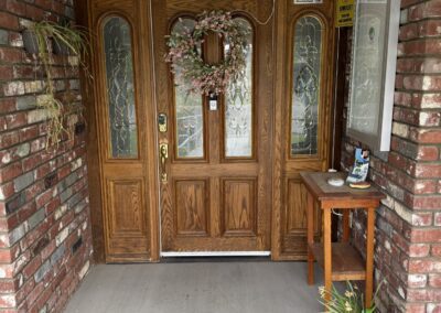 A wooden front door with glass panels, a floral wreath, and a "God Bless this home and all who enter" sign above. A small table with plants and decor sits to the right.