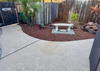 A backyard corner with a curved concrete patio, red mulch, a stone bench, potted plants, and a wooden fence adorned with a metal star.
