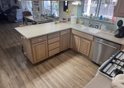 A kitchen with light wood cabinets, a white countertop, a dishwasher, a coffee maker, a white sink, and various items on the counters. The floor has a wood-like finish.
