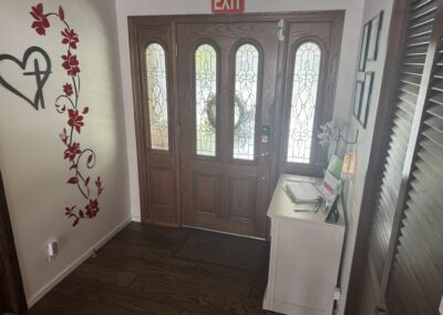 A wooden front door with decorative glass panels, an EXIT sign above, a small white cabinet with papers and flowers, and a wall with red floral decals.