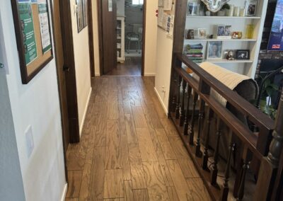 A hallway with wood flooring and framed items on the walls leads to a bathroom. An open railing on the right overlooks a lower room with shelves containing decor and photos.