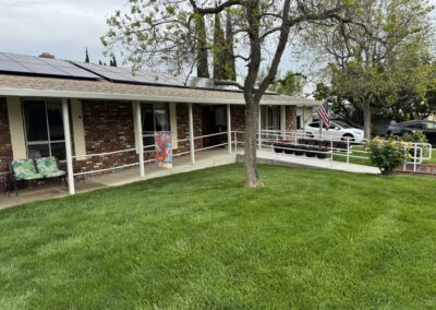 Single-story brick house with a front porch, wheelchair ramp, American flag, green lawn, and trees. Solar panels are visible on the roof.