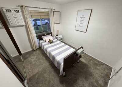 A small bedroom with a single hospital-style bed, striped bedding, a window with beige curtains, framed wall art, and carpeted floor.
