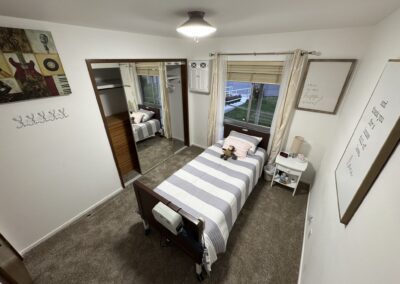 An orderly bedroom with a single bed, striped bedding, a teddy bear, bedside table, lamp, wall art, and mirrored closet doors. Light brown carpet and a window with curtains are visible.