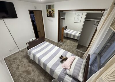 A neatly made single bed with striped bedding, plush toys, and folded clothes in a carpeted bedroom with a mirrored closet and a wall-mounted TV.