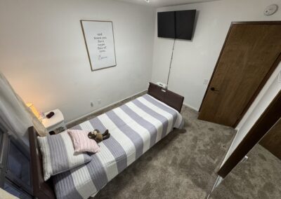 A neatly made twin bed with striped bedding, a teddy bear, and pillows sits in a carpeted bedroom with a wall-mounted TV, wooden door, and framed art above the bed.