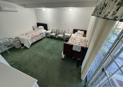A bedroom with two single beds, several chairs, green carpet, floral bedding, a window with curtains, and a medical device at the foot of one bed.